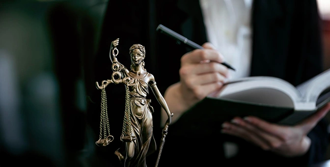 W centrum uwagi znajduje się brązowa statua Lady Justice trzymająca wagę i miecz. W tle osoba pisze w notatniku, nadając całości prawniczy i kontemplacyjny charakter.