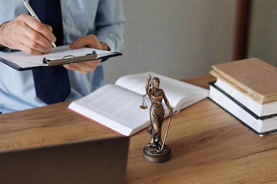 Osoba w niebieskiej koszuli pisze na tabliczce z klipsem, otoczona otwartymi książkami na biurku. Na pierwszym planie stoi mała statuetka Lady Justice, nawiązująca do tematyki prawnej.