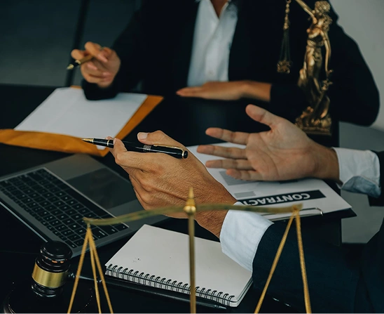 Dwie osoby w strojach biznesowych omawiają dokumenty przy biurku, na którym leżą laptop, umowa, młotek, waga sprawiedliwości i posąg prawnika, nadając rozmowie profesjonalny ton.